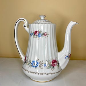 May include: White ceramic coffee pot with a floral design in shades of pink, blue, and green. The pot has a gold trim and a fluted body.