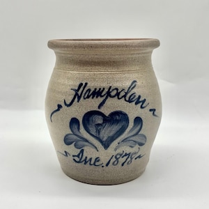 Puede incluir: Un tarro de gres con un diseño de corazón y flores en azul. El tarro tiene la palabra "Hampden" y "Inc. 1878" escritas en escritura azul. El tarro es de color beige claro con un borde más oscuro.
