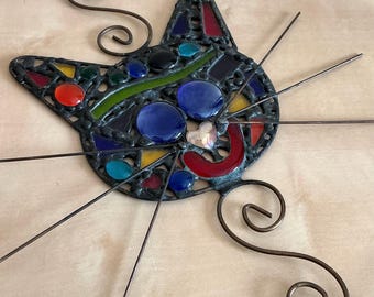Puede incluir: Un rostro de gato de vidrieras con un contorno negro, con coloridas piezas de vidrio. El gato tiene grandes ojos azules, una sonrisa roja y una nariz en forma de corazón. Los acentos de alambre de cobre se extienden desde la cara, y hay espirales decorativas a cada lado.