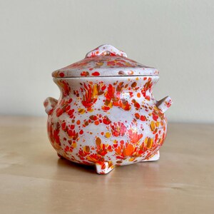 Puede incluir: Un tarro de cerámica con tapa, con un patrón moteado de puntos rojos, naranjas y amarillos sobre un fondo blanco. El tarro tiene dos pequeñas asas y tres patas. La tapa tiene un adorno decorativo.