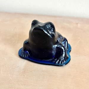 May include: A deep cobalt blue glass frog figurine in a seated position. The translucent glass sculpture features a smooth, rounded body with defined eyes and webbed feet, resting on a flat, integrated base that mimics a small lily pad shape.