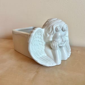 May include: A white ceramic planter with an angel design. The rectangular planter features a detailed relief of an angel with its head resting on its hand and detailed wings. The planter is suitable for small plants or decorative use.