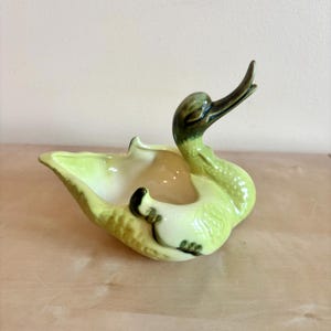 May include: A ceramic swan-shaped planter in shades of green and cream. The swan has a long neck and beak, with a curved body forming a bowl. The planter is likely vintage and suitable for small plants or decorative items.
