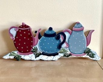 May include: Wooden decorative piece featuring three painted teapots in red, blue, and light blue. Each teapot has white polka dots and is accented with pink. The teapots rest on a white base with green leaf details.