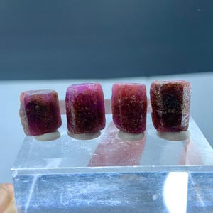 May include: Four rough ruby gemstones, varying in colour from deep red to brownish-red. They are displayed on a clear, rectangular acrylic base, highlighting their natural textures and hues. The background is a soft grey.