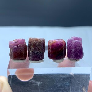 Op de afbeelding: Vier ruwe robijn edelstenen in roze en rode tinten. De natuurlijke robijnen hebben een kristallijne textuur en worden gepresenteerd op een heldere acryl standaard. Deze onbewerkte edelstenen zijn geschikt voor het maken van sieraden of verzamelen.