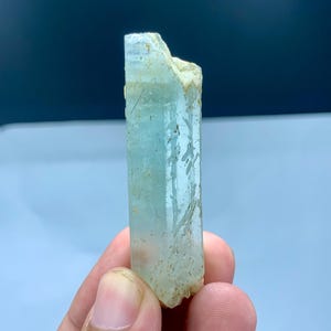 Op de afbeelding: Een ruwe aquamarijnkristal, met een lichtblauwe tint met natuurlijke texturen en onvolkomenheden. Het kristal wordt tegen een zachte blauwe achtergrond gehouden, wat de doorschijnende kwaliteit en natuurlijke vorm benadrukt. Deze edelsteen is een prachtig voorbeeld van natuurlijke minerale formaties.