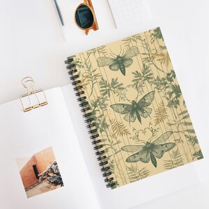 May include: Spiral-bound notebook featuring a beige cover with a botanical design. The cover showcases detailed illustrations of moths and ferns in shades of green and brown. The notebook is open, revealing blank pages.