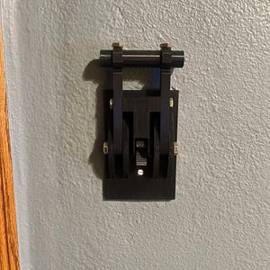 以下が含まれることがあります： 長方形のベースと上部に2本の平行なバーが付いた黒い電気スイッチ。 スイッチは灰色の壁に取り付けられており、小さな金色のネジが付いています。