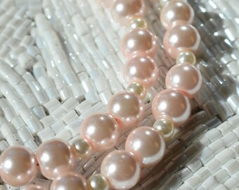 May include: Close-up of two strands of faux pearl necklaces. The larger pearls are a soft pink colour, while the smaller pearls are a creamy white. The necklaces rest on a white, textured surface.