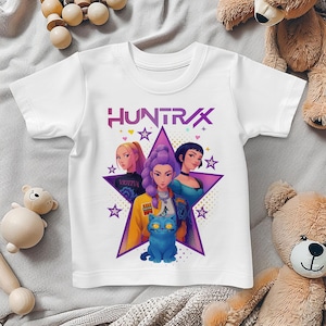 May include: White t-shirt featuring the word "HUNTRAX" in pink and purple. The shirt has a graphic of three women and a blue cat inside a purple star. The design includes stars and hearts.