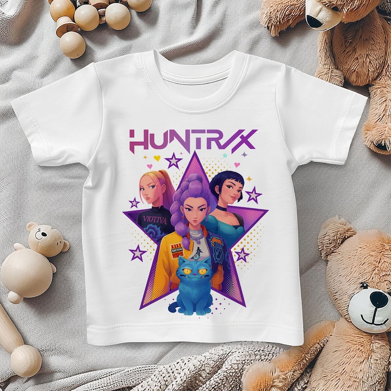 May include: White t-shirt featuring the word "HUNTRAX" in pink and purple. The shirt has a graphic of three women and a blue cat inside a purple star. The design includes stars and hearts.