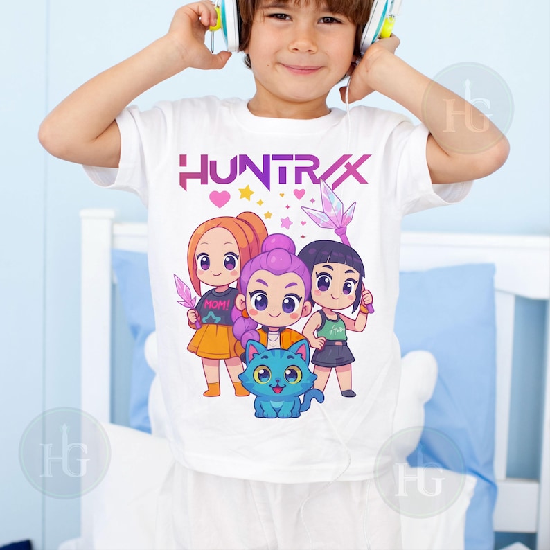 Pu&ograve; includere: T-shirt bianca con la scritta "HUNTRAX" in viola e rosa, e una grafica di tre ragazze dei cartoni animati e un gatto blu. Le ragazze tengono in mano delle armi. La maglietta &egrave; indossata da un bambino.