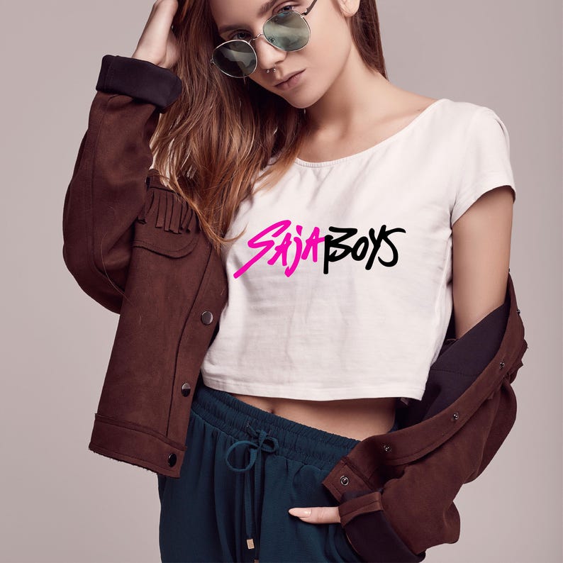 K&ouml;nnte beinhalten: Wei&szlig;es Crop-Top mit dem Text "Saja Boys" in Pink und Schwarz. Das Model tr&auml;gt eine braune Jacke mit Fransen und eine dunkelblaue Hose. Das Model tr&auml;gt auch eine Sonnenbrille.