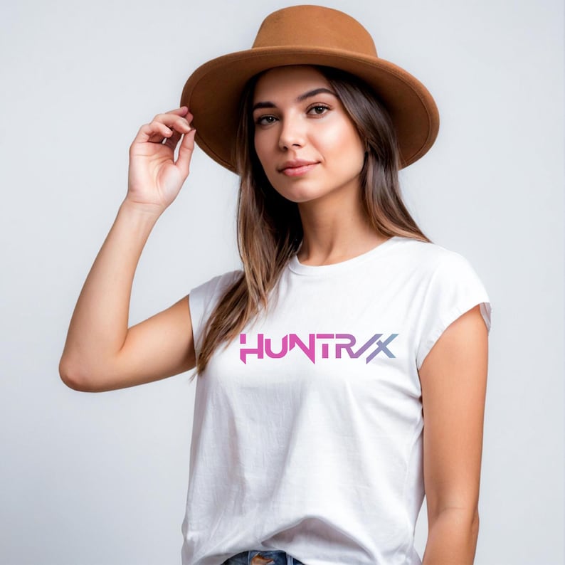 Puede incluir: Camiseta blanca con la palabra "HUNTRX" en degradado rosa y morado. La modelo lleva un sombrero marr&oacute;n. La camiseta tiene cuello redondo y mangas cortas.