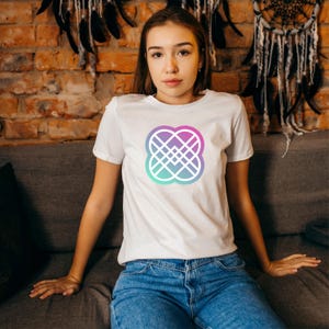 Puede incluir: Camiseta blanca con un dise&ntilde;o geom&eacute;trico en tonos morado, rosa y turquesa. El dise&ntilde;o est&aacute; centrado en la parte delantera de la camiseta. La modelo lleva vaqueros azules.