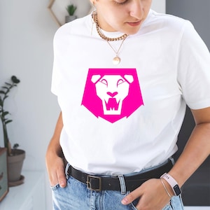K&ouml;nnte beinhalten: Wei&szlig;es T-Shirt mit einem leuchtend pinkfarbenen L&ouml;wenkopf-Motiv. Das L&ouml;wen-Design ist geometrisch und k&uuml;hn, in einer Schildform. Das Shirt ist aus weichem Material und wird mit blauen Jeans und einem schwarzen G&uuml;rtel getragen.