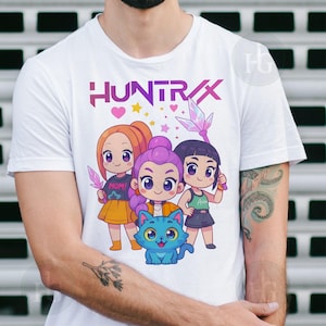 Pu&ograve; includere: T-shirt bianca con la scritta "HUNTRAX" in viola e rosa, e un'illustrazione a cartoni animati di tre ragazze e un gatto blu. Le ragazze tengono in mano dei cristalli e indossano abiti diversi.