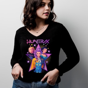 May include: Black long-sleeve V-neck shirt featuring a graphic of three women and a blue cat inside a star. The word "HUNTRAX" is above the star. The women have different hair colors and wear various outfits.