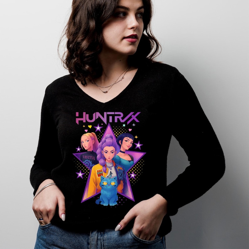 May include: Black long-sleeve V-neck shirt featuring a graphic of three women and a blue cat inside a star. The word "HUNTRAX" is above the star. The women have different hair colors and wear various outfits.