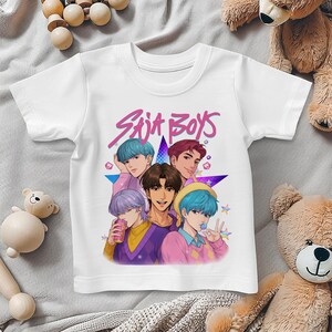 May include: White t-shirt featuring a colorful graphic of five anime-style characters with different hair colors. The text "Saja Boys" is written in pink above the characters. The shirt is made of a soft material.