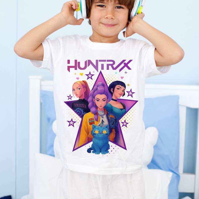 May include: White t-shirt featuring a graphic of three stylized women inside a purple star, with a blue cat at the bottom. The word "HUNTRAX" is printed above the star in purple and pink. Additional stars surround the main graphic.