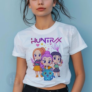 Pu&ograve; includere: T-shirt bianca con la scritta "HUNTRAX" in viola e una grafica a cartoni animati di tre ragazze e un gatto blu. Le ragazze tengono in mano dei cristalli e indossano abiti colorati. La maglietta &egrave; girocollo.