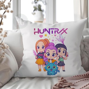 Pu&ograve; includere: Cuscino decorativo bianco con una grafica a cartoni animati colorata. Il design include tre figure stilizzate, un gatto blu e la scritta "HUNTRAX" in viola. Le figure tengono in mano oggetti simili a cristalli.