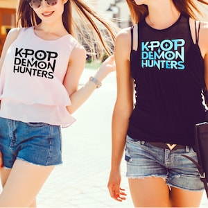 K&ouml;nnte beinhalten: Zwei Frauen tragen &auml;rmellose Oberteile und Jeansshorts. Ein Oberteil ist hellrosa mit dem Text "KPOP DEMON HUNTERS" in Schwarz. Das andere Oberteil ist schwarz mit dem gleichen Text in Blau.
