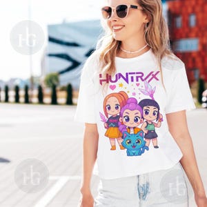 Pu&ograve; includere: T-shirt bianca con la scritta "HUNTRAX" e un'illustrazione di tre ragazze dei cartoni animati e un gatto blu. Le ragazze hanno colori di capelli diversi e tengono in mano oggetti. Abbigliamento casual.
