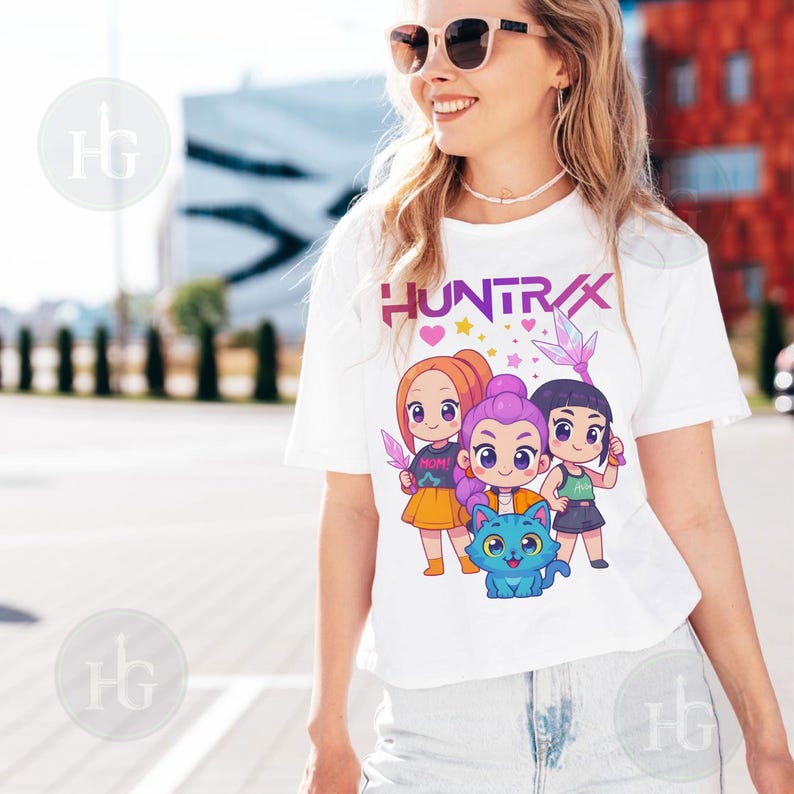 Pu&ograve; includere: T-shirt bianca con la scritta "HUNTRAX" e un'illustrazione di tre ragazze dei cartoni animati e un gatto blu. Le ragazze hanno colori di capelli diversi e tengono in mano oggetti. Abbigliamento casual.