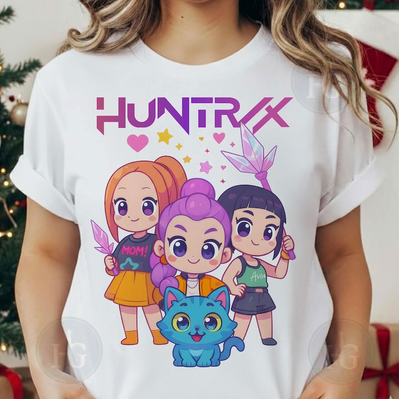 Pu&ograve; includere: T-shirt bianca con una grafica colorata di tre ragazze dei cartoni animati e un gatto blu. La parola "HUNTRAX" &egrave; stampata sopra i personaggi. Le ragazze tengono in mano oggetti a forma di cristallo. La maglietta di una ragazza dice "MOM!" e un'altra "Ava".