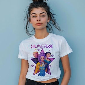 May include: White t-shirt featuring the word "HUNTRAX" in purple, with a star graphic containing illustrations of three women and a blue cat. The shirt has short sleeves and a crew neck.