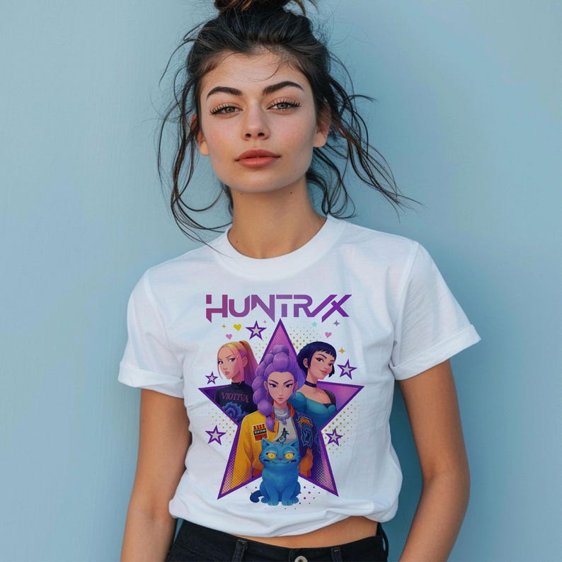 May include: White t-shirt featuring the word "HUNTRAX" in purple, with a star graphic containing illustrations of three women and a blue cat. The shirt has short sleeves and a crew neck.