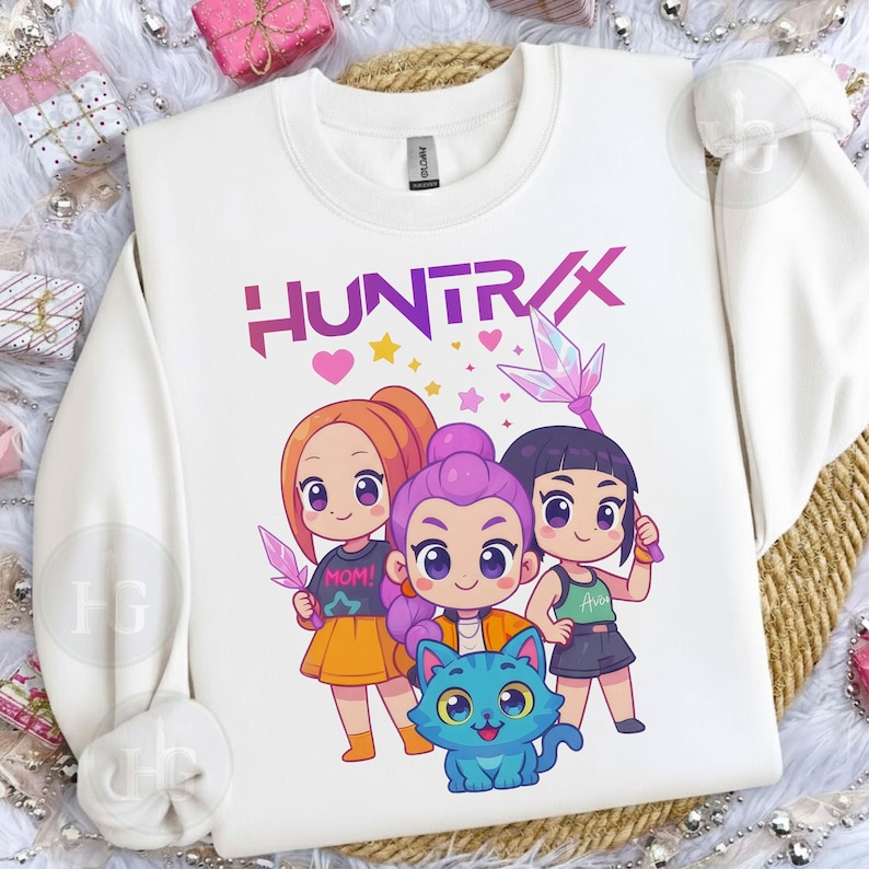 Pu&ograve; includere: Felpa bianca con una grafica colorata di tre ragazze dei cartoni animati e un gatto blu. La parola "HUNTRIX" &egrave; stampata sopra i personaggi in viola. Le ragazze tengono in mano oggetti simili a cristalli.