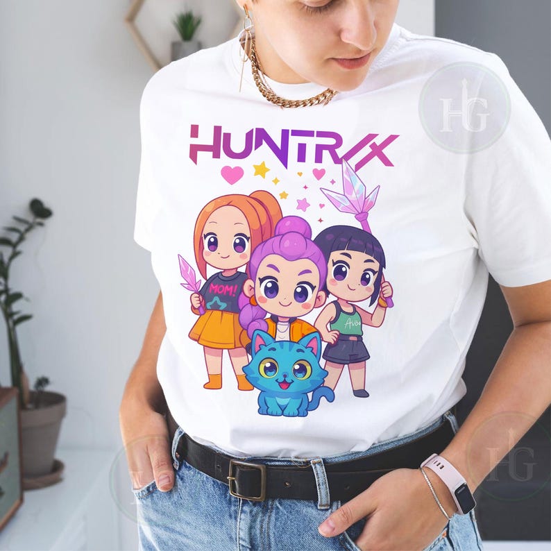 Pu&ograve; includere: T-shirt bianca con una grafica colorata di tre ragazze dei cartoni animati e un gatto blu. La parola "HUNTRIX" &egrave; stampata sopra i personaggi in viola e rosa. Le ragazze tengono in mano oggetti simili a cristalli.