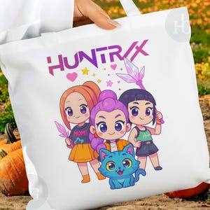 Pu&ograve; includere: Borsa tote bianca con la scritta "HUNTRIX" in rosa e viola. La borsa presenta un'illustrazione a cartoni animati di tre ragazze e un gatto blu. Una ragazza indossa una maglietta con la scritta "MOM!"