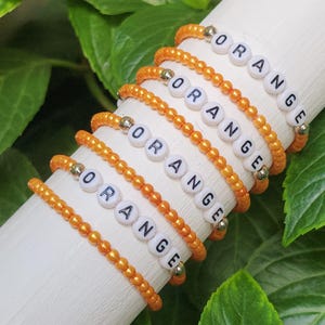 May include: Several orange beaded bracelets with the word "ORANGE" spelled out in white letter beads. The bracelets are displayed on a white cylindrical surface, with green leaves in the background.