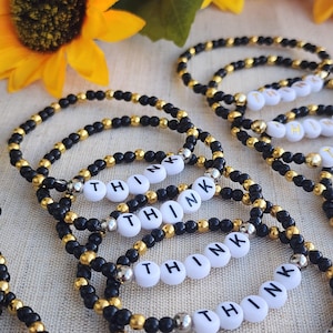 Pode incluir: Várias pulseiras de contas com a palavra "THINK" escrita em contas de letras brancas. As pulseiras apresentam contas pretas e douradas, com algumas contas prateadas separando as contas de palavras. O fundo inclui um girassol e um tecido neutro.