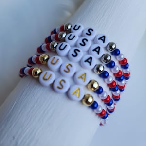 May include: A stack of beaded bracelets in red, white, and blue, with the letters "USA" spelled out on white beads. Some beads are gold or silver. The bracelets are on a white surface.
