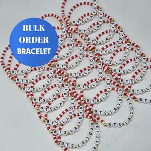 Pode incluir: Uma coleção de pulseiras de contas com contas vermelhas e brancas alternadas, pequenas contas espaçadoras douradas e contas de letras brancas que soletram BTS LA 2026. Um gráfico circular azul exibe o texto branco BULK ORDER BRACELET.