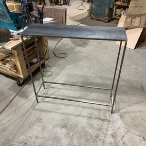 May include: A metal console table with a rectangular top and a lower shelf. The table has a dark, weathered finish and a minimalist design. The legs and frame are made of thin metal rods.