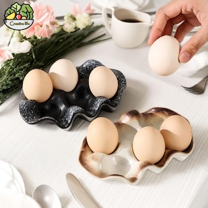 May include: Two ceramic egg holders, one black and one brown, each holding several brown eggs. The black holder has a speckled design, while the brown one has a wavy edge. A hand holds an egg above the holders.
