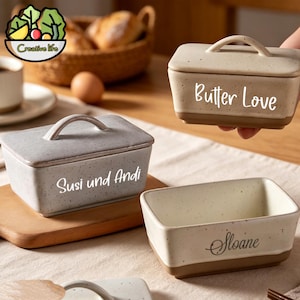 May include: Three rectangular ceramic butter dishes with lids and handles. One cream-coloured dish is held, labelled Butter Love. A grey dish is labelled Susi und Andi. A cream-coloured open dish is labelled Sloane. All feature a speckled finish and brown base.