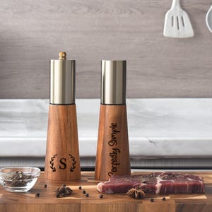 May include: Two wooden pepper grinders with stainless steel tops. One grinder has the letter "S" inside a laurel wreath, the other has the words "Perfectly Simple". A small glass bowl of peppercorns and a piece of raw meat are also visible.