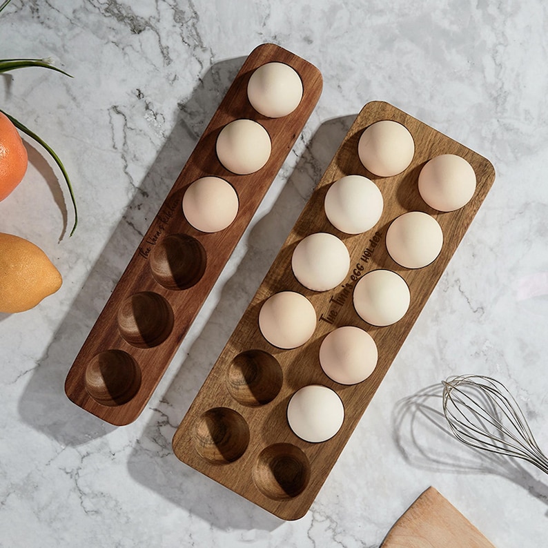 K&ouml;nnte beinhalten: Zwei Eierhalter aus Holz, einer mit dem Text "The Hive" und der andere mit dem Text "True Thyme Egg Holder". Beide Halter sind mit wei&szlig;en Eiern gef&uuml;llt.