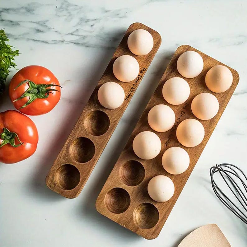 K&ouml;nnte beinhalten: Zwei h&ouml;lzerne Eierhalter mit dem Text "The Egg's Kitchen" darauf. Die Halter sind mit wei&szlig;en Eiern gef&uuml;llt.
