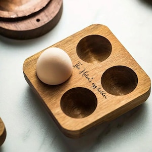 K&ouml;nnte beinhalten: Ein Eierhalter aus Holz mit vier F&auml;chern. Der Halter ist mit dem Text "The Helen's egg holder" graviert. Ein Ei befindet sich im Halter.