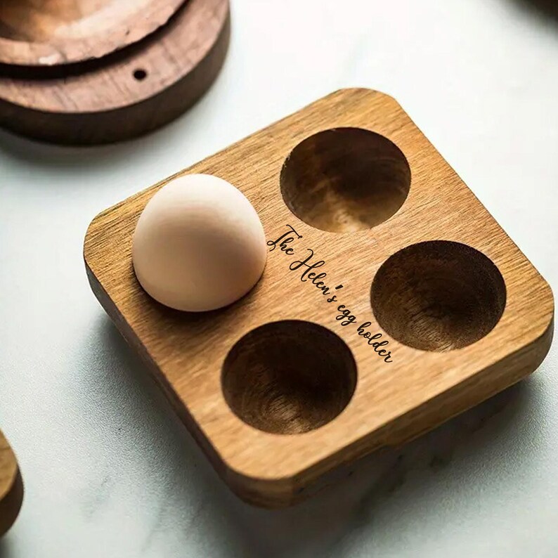 K&ouml;nnte beinhalten: Ein Eierhalter aus Holz mit vier F&auml;chern. Der Halter ist mit dem Text "The Helen's egg holder" graviert. Ein Ei befindet sich im Halter.