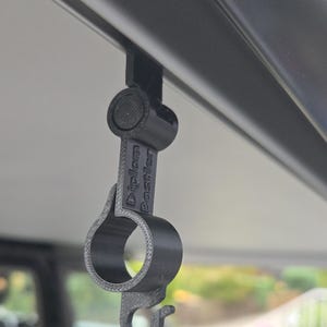 Puede incluir: Un clip negro impreso en 3D con el texto "Diplom Bastler", diseñado para colgar del borde de un vehículo. El clip tiene una abertura circular y forma de gancho.